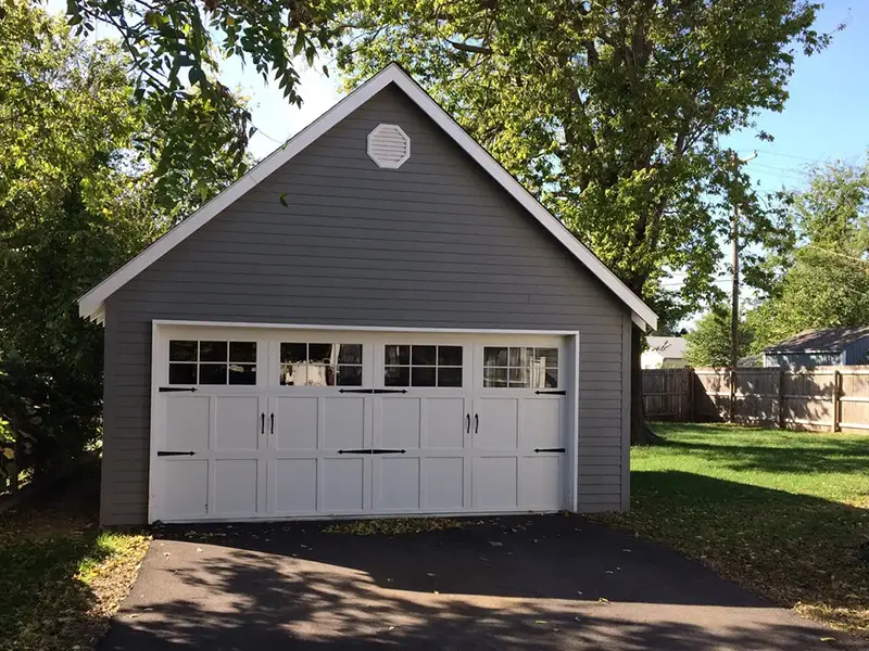 A gray garage with two white doors and a tree in the background.